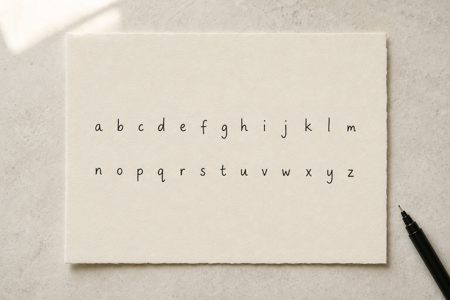 A cream sheet of paper on a soft stone desk showing a full lowercase alphabet, a to z, written by hand in black fineliner with even spacing between each letter.